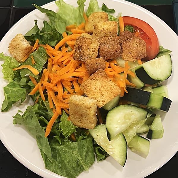 A side salad with lettuce, shredded carrots, croutons, cucumber slices, and a tomato wedge.