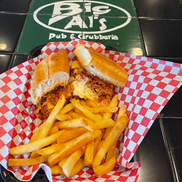 Meatball sub and fries in a basket with red and white checkered paper, Big Al's logo in background.