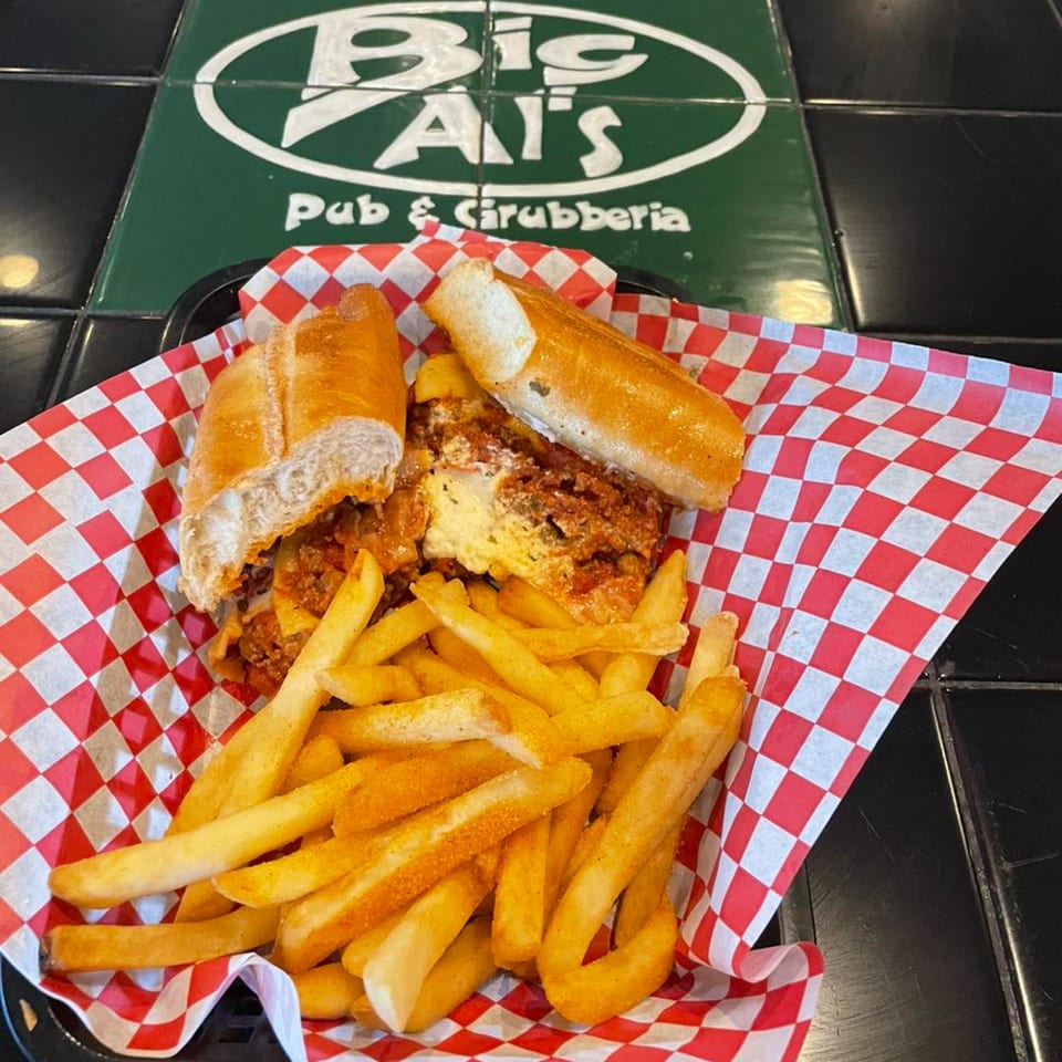 Meatball sub and fries in a basket with red and white checkered paper, Big Al's logo in background.