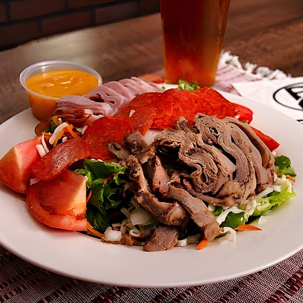 A large chef salad with various meats, tomatoes, and lettuce, with a side of dressing and a beer.