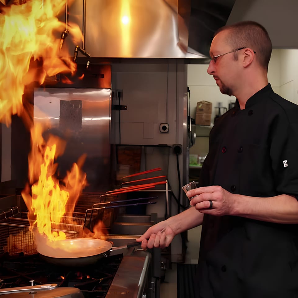 A chef in a black uniform flambés food in a pan with large flames in a commercial kitchen.