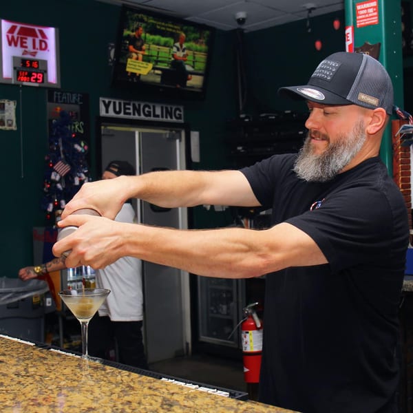 Bartender shaking a cocktail at Big Al's Pub & Grubberia, with a TV screen in the background.